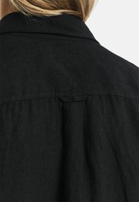 Zwarte linnen shirt met een rugplooi, subtiele textuur en een gestructureerde kraag. De stof lijkt licht en ademend te zijn.