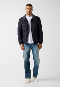 Marineblaue Daunenjacke mit gestepptem Design, grauem Rippkragen, kombiniert mit hellblauen, verwaschenen Jeans und weißen Turnschuhen.