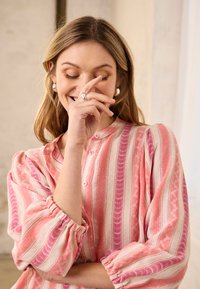 Moteris su užmerktomis akimis šypsosi, ranka iš dalies dengia veidą, dėvi sidabrinius auskarus ir rožinę dryžuotą ilgomis rankovėmis palaidinę.