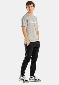 Graues Baumwoll-T-Shirt mit weißem Logo, schwarze Hose mit Seitentaschen und schwarz-weiße Sneakers mit gelben Akzenten, stehend vor einem weißen Hintergrund.
