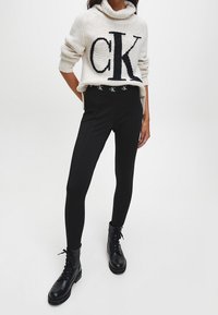 Jasno beżowy sweter z dużym czarnym logo "CK", zestawiony z dopasowanymi czarnymi spodniami z logo w pasie i czarnymi butami wojskowymi.