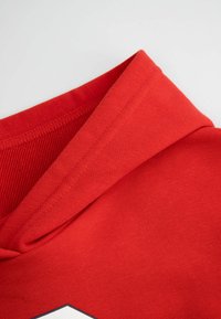 Czerwona bluza z kapturem z teksturowanym kapturem, gładkim materiałem i kontrastującym białym graficznym detalem z przodu. Czyste szwy wokół krawędzi.