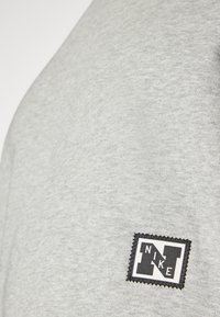 Helle graue Sweatshirtstoffe mit einem strukturierten, gestickten Schwarz-Weiß-Nike-Logo-Patch im linken Brustbereich.