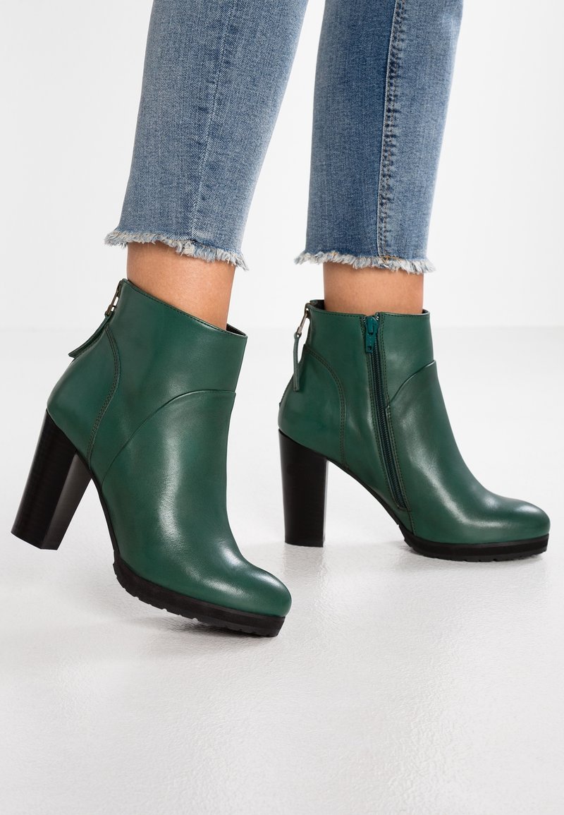 Bottes à chevilles en cuir vert avec talons noirs épais, fermetures éclair latérales, portées avec un jean bleu à ourlet effiloché sur un sol blanc.