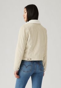Giacca in corduroy beige con colletto in shearling bianco, caratterizzata da un design corto e chiusure a bottoni. Abbinata a jeans in denim blu.