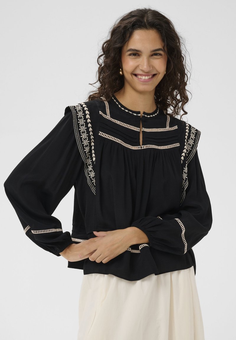 Femme souriante aux cheveux bouclés portant un chemisier noir avec broderies blanches et une jupe crème, debout devant un fond blanc.