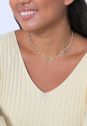 Collar de cuentas colorido que presenta pequeñas cuentas en tonos pastel con un charm central de cara sonriente amarilla, sobre una blusa texturizada de color amarillo claro.