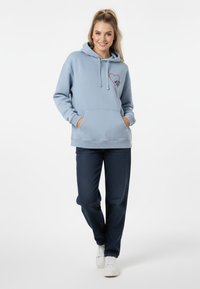 Felpa azzurro chiaro con tasca a marsupio, decorata con una grafica di un cuore rosso; abbinata a jeans scuri e sneakers bianche. Tessuto liscio e texture.