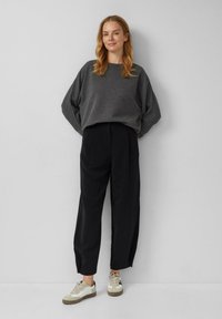 Femme aux cheveux châtain clair portant un pull ample gris foncé, un pantalon large noir et des baskets blanches, se tenant contre un mur blanc.