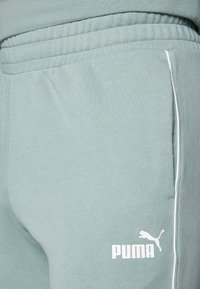 Pantalon de survêtement vert clair avec une texture douce, présentant des rayures blanches sur les côtés et un logo Puma blanc en bas à gauche de la jambe. Ceinture élastique incluse.
