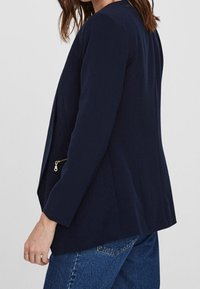 Tmavě modrý blazer s dlouhými rukávy a detaily na zipu. Hladká tkanina, strukturovaný design a mírně rozšířený tvar. Nosený s modrými džínami.
