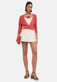 Koraalkleurige cardigan over een witte v-hals top, gecombineerd met een witte rok. Getextureerde slangenhuid hakken en een bijpassende rode handtas. Geometrische ketting.