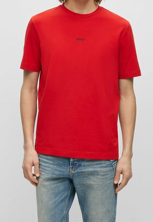 Personne portant un t-shirt rouge vif à manches courtes avec le texte "BOSS" sur la poitrine et un jean bleu clair sur un fond uni.