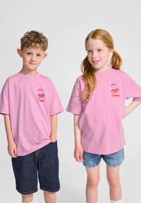 Rosa t-shirts med kort ärm och körsbärsgrafik samt logotyp, bärs av två barn. Ett barn har på sig jeansshorts, det andra har ljusblå shorts.