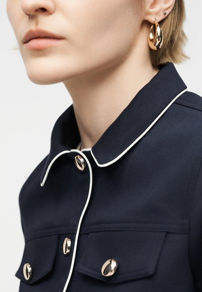 Κοντινό πλάνο ενός ατόμου που φοράει μπλε ναυτικό μπουφάν με λευκή διακοσμητική ραφή και χρυσά κουμπιά, καθώς και μεγάλα χρυσά κρίκους σκουλαρίκια με δύο μικρότερα σκουλαρίκια επάνω.