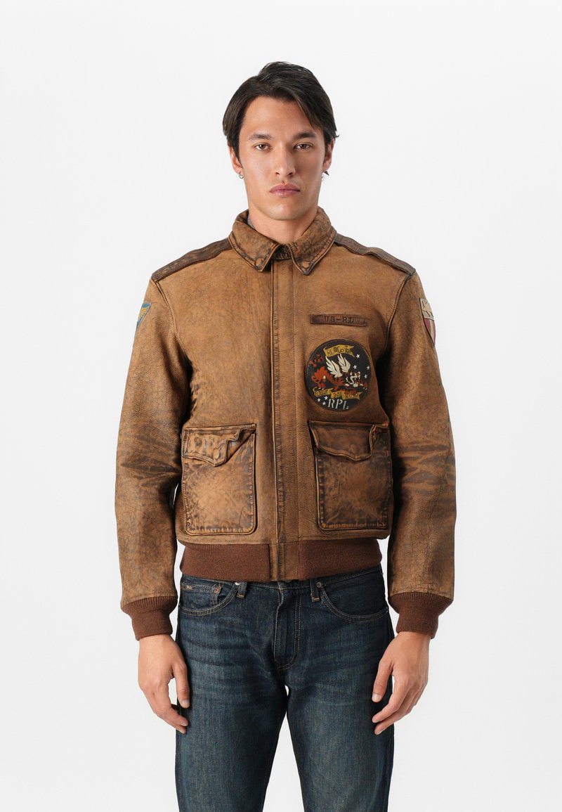 Giacca bomber in pelle marrone con zip frontale, quattro tasche e toppe ricamate; polsini e orlo a costine; finitura liscia e tessurizzata.