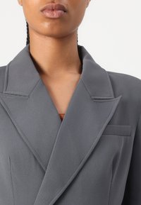 Femme portant un blazer gris tailleur avec de larges revers et une poche poitrine visible sur fond blanc.