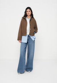 Καφέ κοντή ζακέτα με κουμπιά πάνω από ένα γαλάζιο ριγέ πουκάμισο, συνδυασμένη με φαρδιά μπλε τζιν και λευκές γόβες, σε ουδέτερο φόντο.