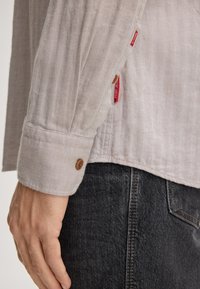 Camicia a maniche lunghe a spina di pesce grigia chiara con polsini abbottonati, dotata di etichette rosse con logo. Abbinata a pantaloni in denim scuri, che mostrano una trama testurizzata.