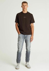 Bruine katoenen t-shirt met korte mouwen en een subtiel logo, gecombineerd met lichtblauwe denim jeans en grijze sneakers, staand op een witte achtergrond.