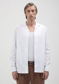 Uomo che indossa una camicia bianca a bottoni aperta sopra una canotta bianca, pantaloni marroni e una cintura nera, in piedi contro uno sfondo grigio chiaro uniforme.