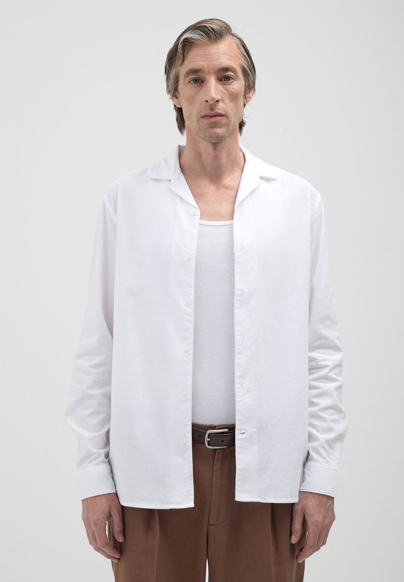 Uomo che indossa una camicia bianca a bottoni aperta sopra una canotta bianca, pantaloni marroni e una cintura nera, in piedi contro uno sfondo grigio chiaro uniforme.