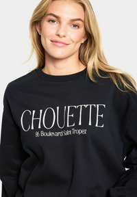 Čierna mikina vyrobená z bavlny, s bielym potlačeným textom "CHOUETTE 86 Boulevard Saint Tropez" v tučnom písme na prednej strane.