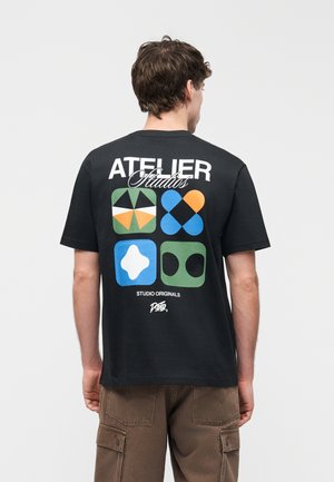 Mand iført sort t-shirt med farverige geometriske former og teksten "Atelier Studios" på ryggen, kombineret med brune cargobukser.
