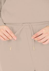 Beige loungewear met een zachte textuur, voorzien van een tailleband met een trekkoord en gouden metalen uiteinden. Handen die de tailleband vasthouden voor aanpassing.