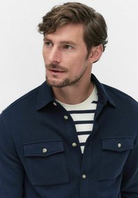 Marinblå skjorta med knappar och två bröstfickor, strukturerad tygkvalitet och silverfärgade knappar. Underneath syns en vit skjorta med marinblå ränder.