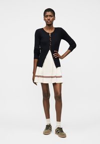 Femme portant un cardigan noir boutonné, jupe plissée blanche avec des rayures rouges, chaussettes beiges et baskets vert olive, debout avec la main sur la hanche.