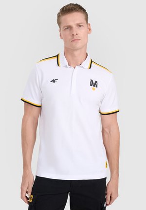 Mężczyzna w białej koszulce polo z czarnymi i żółtymi wykończeniami oraz logo, stoi na tle jednolitego jasnoszarego tła.