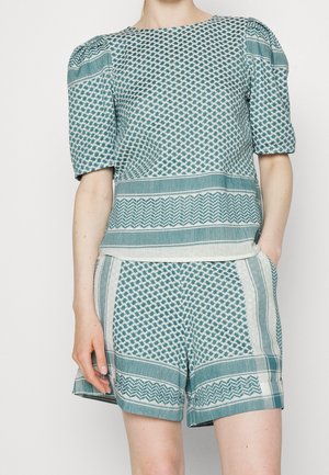 Vrouw draagt een kort mouw bovenstuk met opgestikte schouders in teal en wit patroon en bijpassende korte broek, handen in de zakken.