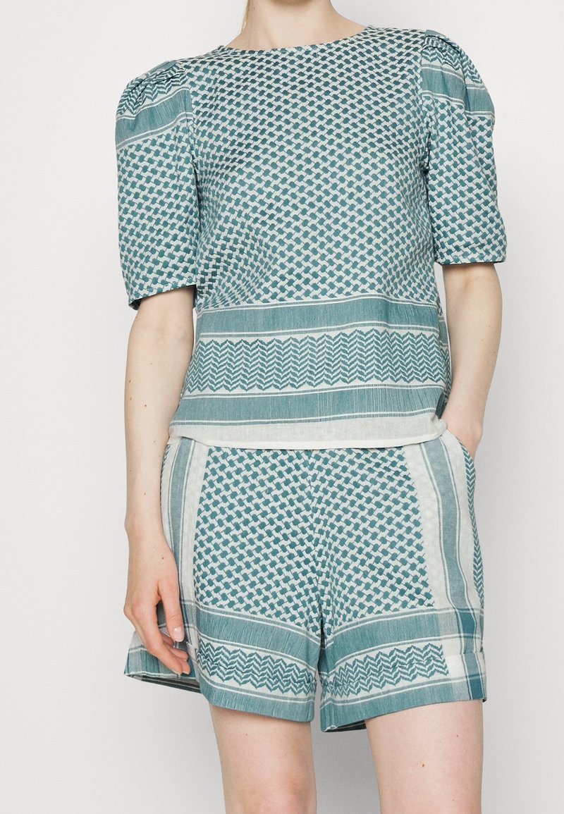 Femme portant un haut à manches courtes bleu sarcelle et blanc à motifs avec épaules bouffantes et short assorti, les mains dans les poches.