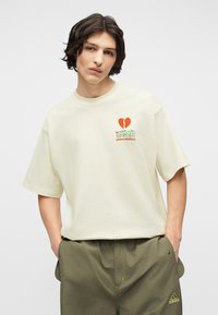 Νεαρός άντρας με σκουρόχρωμα μαλλιά φοράει εκρού μπλουζάκι Adidas με κόκκινη σπασμένη καρδιά και ιαπωνικό κείμενο, συνδυασμένο με χακί παντελόνι Adidas.