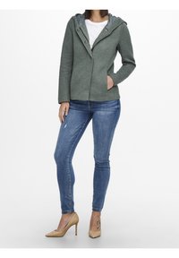 Grön huva jacka med texturerad yta och knappstängning, kombinerad med åtsittande blå jeans och beiga klackar. Synlig mönstrad tatuering på vristen.