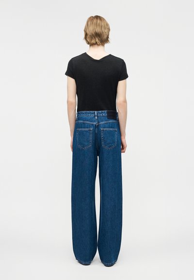 Άτομο με κοντά ξανθά μαλλιά που φοράει μαύρο μπλουζάκι και ψηλόμεσα φαρδιά μπλε τζιν παντελόνια, στέκεται με την πλάτη προς τα εμπρός πάνω σε απλό φόντο.