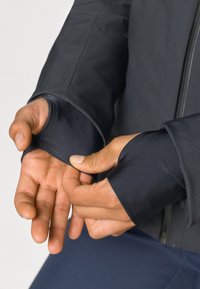 Giacca sportiva nera con fori per i pollici nei polsini. Tessuto liscio e elastico, design aderente, che mostra le mani in una posizione rilassata.