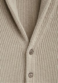 Beige knit cardigan featuring a V-neck, ribbed texture, and three brown buttons along the front. Soft, chunky material with a cozy appearance.