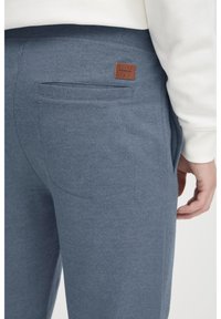 Pantalones de chándal grises confeccionados en una tela suave, con un bolsillo trasero y un parche de etiqueta de cuero marrón. Cintura elástica con un acabado suave.