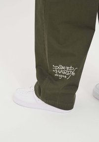 Olijfgroene broeken met een losse fit, voorzien van witte geborduurde tekst "Don Ed Hardy designs" en gedragen met witte sneakers.
