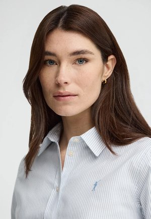 Mujer con cabello largo y castaño, lleva pendientes pequeños de aro dorado y una camisa de botones a rayas azul claro con un pequeño logo bordado.