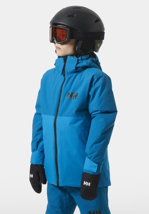 Kind in blauer Skijacke und Skihose, trägt schwarzen Helm mit orangefarbener Skibrille und schwarze Handschuhe, steht und schaut nach links.