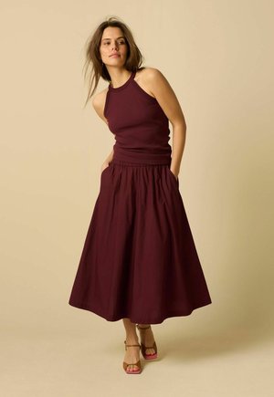 Vrouw met een mouwloze bordeaux top en bijpassende lange rok, staand met handen in de zakken tegen een beige achtergrond.