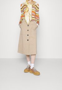 Jupe midi beige avec des boutons à l'avant, associée à une chemise rayée à manches longues et des chaussures à lacets épaisses en marron avec une semelle en caoutchouc.