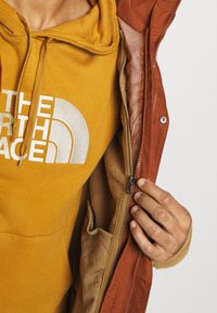 Žlutá mikina s bílým logem pod hnědou bundou, která má fleecovou podšívku, zapínání na zip a viditelnou vnitřní kapsu.