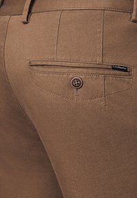 Brązowe spodnie o teksturowej powierzchni, z zapiętą kieszenią na plecach i małą metką marki, zaprojektowane do użytku codziennego.