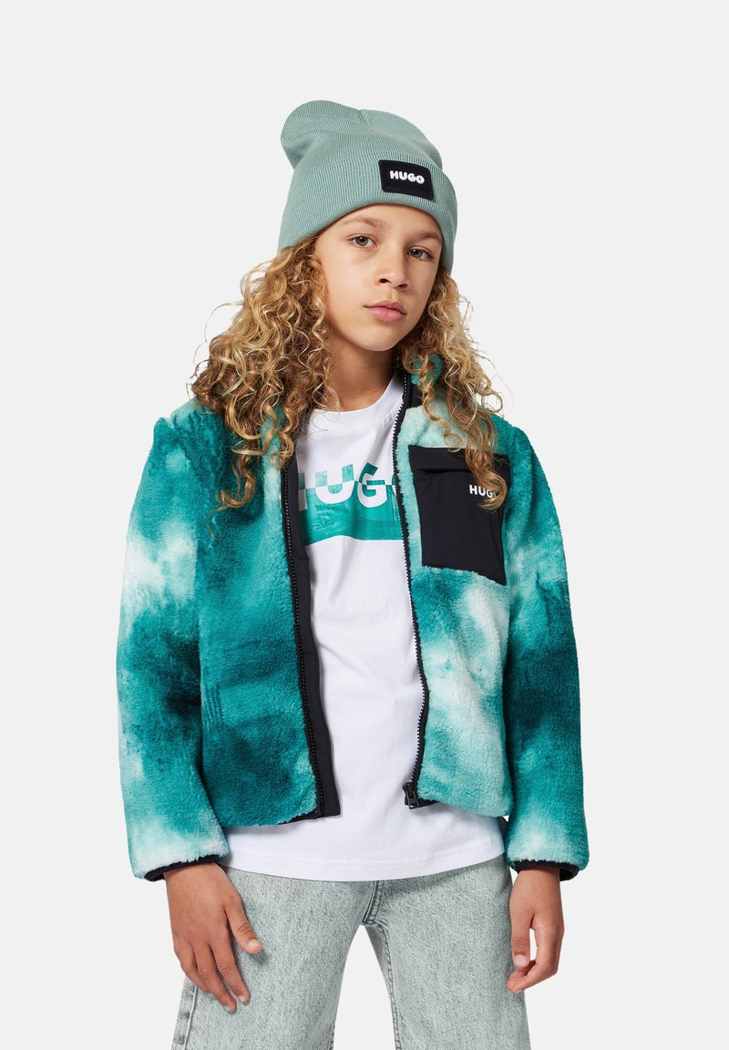 Chaqueta de piel sintética en verde azulado y negro con cierre de cremallera, combinada con una camiseta blanca con un logo gráfico y jeans de color gris claro.