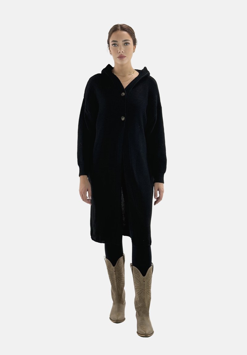 Svart stickad lång cardigan med huva, med knapplåsning och ribbad textur, bärs över svarta leggings och cowboyboots.