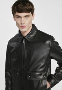 Czarna skórzana kurtka z kołnierzem, z przodu na zamek, dwiema naszytymi kieszeniami i gładką fakturą, z widocznymi szwami i zatrzaskami.